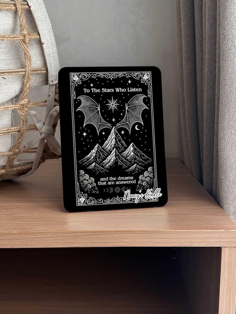 K&ouml;nnte beinhalten: Schwarze rechteckige Kindle-H&uuml;lle mit wei&szlig;en Illustrationen von Bergen, Flederm&auml;usen, Sternen und Monden. Der Text lautet "To The Stars Who Listen" und "and the dreams that are answered". Die H&uuml;lle hat einen dekorativen Rand und den Namen "Marcy's Kindle".