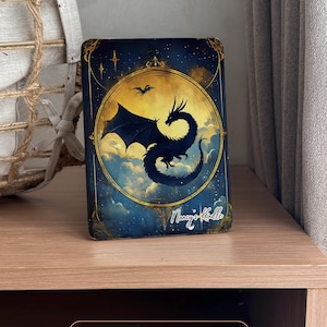 May include: A dark blue and gold illustrated card with a black silhouette of a dragon flying in a circle with clouds and stars. The card has the text "Nancy's Kindle" in the bottom right corner.