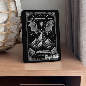 K&ouml;nnte beinhalten: Schwarze E-Reader-H&uuml;lle mit einem Himmelsdesign. Es zeigt Berge, Sterne und fledermausartige Fl&uuml;gel. Der Text lautet "To The Stars Who Listen" und "and the dreams that are answered". Personalisiert mit "Nancy's Kindle".