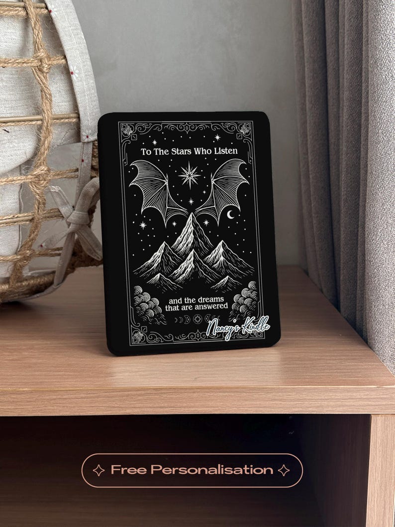 K&ouml;nnte beinhalten: Schwarze E-Reader-H&uuml;lle mit einem Himmelsdesign. Es zeigt Berge, Sterne und fledermausartige Fl&uuml;gel. Der Text lautet "To The Stars Who Listen" und "and the dreams that are answered". Personalisiert mit "Nancy's Kindle".