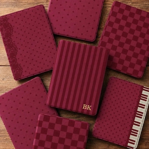 May include: A collection of six rectangular covers in shades of deep burgundy. The designs include polka dots, a checkered pattern, vertical stripes with the gold letters BK, and a piano key motif along the edge of one cover with lace accents.