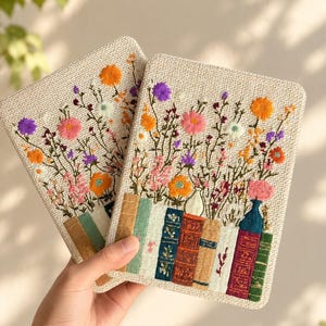 May include: Two rectangular fabric covers featuring detailed embroidery of a row of colourful books at the base with a vibrant bouquet of wildflowers in shades of orange, pink, purple, and white extending upwards from the book spines.