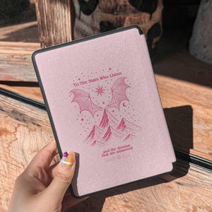 May include: A pink e-reader cover with a black border. The cover features a design with the text "To The Stars Who Listen" and "and the dreams that are answered". The design includes illustrations of mountains, wings, and a star.
