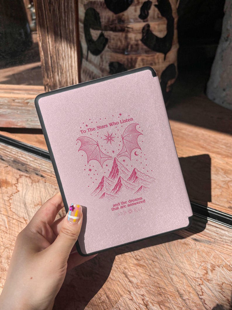 May include: A pink e-reader cover with a black border. The cover features a design with the text "To The Stars Who Listen" and "and the dreams that are answered". The design includes illustrations of mountains, wings, and a star.