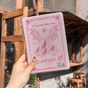 May include: A pink rectangular case with the text "To The Stars Who Listen" and "and the dreams that are answered". The case features a mountain and wing design. The name "Thida" is printed on the bottom right.