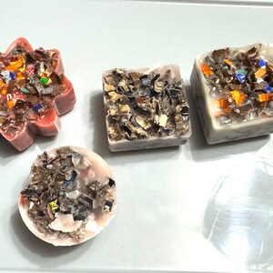 May include: Four handmade candles with different shapes and colors. The candles are made with embedded pieces of paper and other materials. The candles are on a white surface.