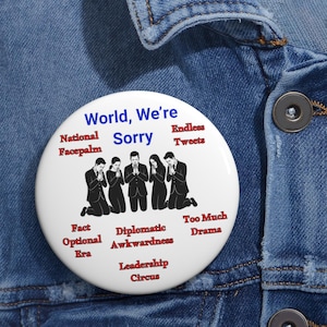 Può includere: Un bottone bianco con la scritta "World, We're Sorry" in blu, e altre frasi in rosso. Presenta una grafica di persone inginocchiate in preghiera. Il bottone è appuntato su una giacca di jeans.