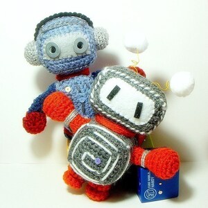 May include: Two crocheted robot toys, one blue and gray with a black headband and the other gray and red with a white face. The robots are holding each other and standing on a blue box with the text "CUBE WORLD HANDY" printed on it.