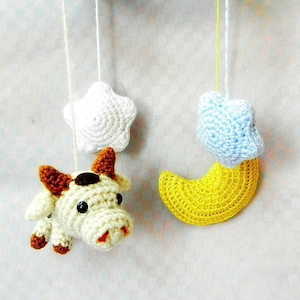 Puede incluir: Un móvil de bebé de crochet con una estrella blanca, una estrella azul, una luna creciente amarilla y una vaca marrón y blanca.