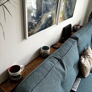 May include: A long, narrow wooden console table positioned behind a teal fabric sofa. The table holds two ceramic mugs with colourful illustrations and a remote control rests on the sofa cushion. Two framed abstract paintings hang on the wall above.