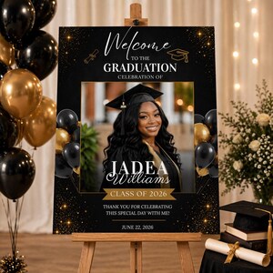 Puede incluir: Un letrero de bienvenida de graduación con el texto "Bienvenida a la celebración de graduación de JADEA Williams, Clase de 2026". El letrero es negro con detalles dorados y muestra una foto de la graduada. Globos negros y dorados en el fondo.
