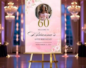 May include: A pink and gold 60th birthday welcome sign on a gold easel. The sign features a photo of a woman, the number 60, and the text "Welcome to Brianna's 60th Birthday November 21, 2025". The sign also includes floral accents.
