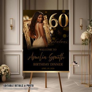 May include: A black and gold birthday welcome sign on a wooden easel. The sign features a photo of a woman in a gold dress, the number 60, and the words "Welcome to Amelia Smith Birthday Dinner April 20, 2026."