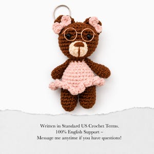 Puede incluir: Un llavero de oso de ganchillo hecho a mano en tonos marrón y rosa. El oso lleva un vestido rosa y gafas redondas pequeñas. El llavero tiene un anillo plateado en la parte superior. El texto de la imagen dice: "Written in Standard US Crochet Terms. 100% English Support - Message me anytime if you have questions!"