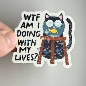 Peut inclure: Autocollant holographique représentant un chat noir avec une tête en forme de bocal à poissons contenant un poisson jaune. Le texte "WTF AM I DOING WITH MY LIVES?" est inclus. Le chat est assis sur un tabouret en bois avec un fond étoilé.