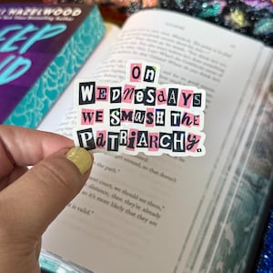 May include: A sticker with the text "On Wednesdays We Smash the Patriarchy" in a black, pink, and white design. The sticker is held over an open book.