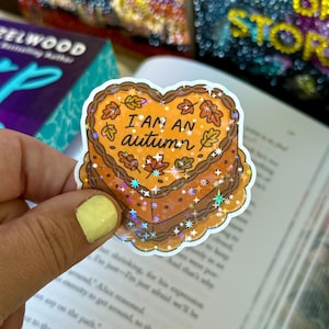 May include: A heart-shaped sticker with the text "I AM AN autumn" in black. The sticker has a brown cake design with orange leaves and glitter accents. The sticker is held in a hand.