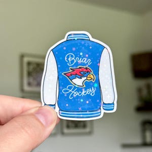 May include: A sticker in the shape of a blue and white varsity jacket. The jacket has white sleeves and a blue body with the words "Briar Hockey" and a bird logo. The sticker is held by a hand.