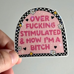 May include: A pink sticker with the text "OVER FUCKING STIMULATED & NOW I'M A BITCH" in pink. The sticker has a black and white checkered border, with small flower and star accents.