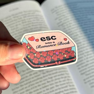 May include: A sticker with the text "esc into a Romance Book" in black and pink. The sticker is shaped like a box filled with red roses. Two pink hearts are above the word "esc". The sticker has a holographic, glittery effect.