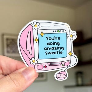 May include: A pink and white sticker featuring a retro computer design with the words "You're doing amazing sweetie" on the screen. The sticker is decorated with yellow stars and white daisy flowers.