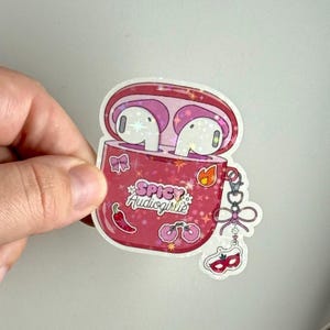 Może przedstawiać: Różowa naklejka przedstawiająca otwarte etui na AirPods z dwoma białymi słuchawkami w środku. Etui zawiera napis "Spicy Audiogirls" oraz elementy dekoracyjne, takie jak papryczka chili, wiśnie i płomień.