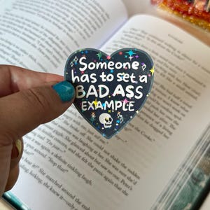 以下が含まれることがあります： 「Someone has to set a BAD ASS EXAMPLE」の白い文字が暗い青色の背景に書かれたハート型のピン。ピンには、ドクロと星のデザインがあります。開いた本の上にピンが置かれています。