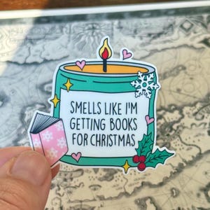 May include: A sticker featuring a teal candle with a burning flame, snowflake, and hearts. The text reads "Smells like I'm getting books for Christmas." A small pink book with snowflakes is also included.