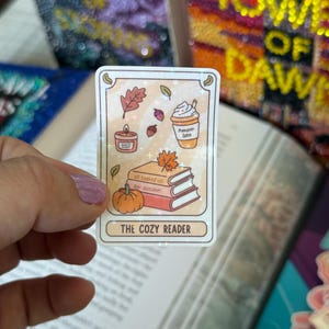 May include: A rectangular sticker with a fall theme. The sticker features illustrations of a pumpkin spice latte, a candle, leaves, acorns, a pumpkin, and books. The text "all booked up for autumn" and "THE COZY READER" are also included.