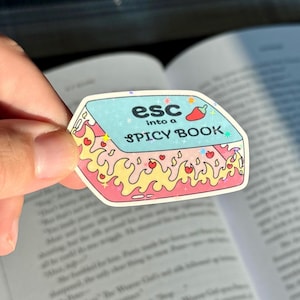 May include: A sticker with the text "esc into a SPICY BOOK" on a light blue background. The sticker has a book design with flames and hearts around the edges and a red chili pepper icon.