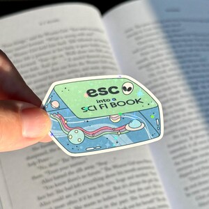 May include: A holographic sticker with the text "esc into a SCI FI BOOK" in green and white. The sticker features a design of a spaceship, planets, and a colorful wormhole. The sticker is held over an open book.