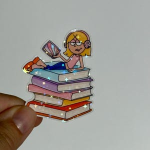 Può includere: Un adesivo con un'illustrazione a cartoni animati di una persona che legge, sdraiata su una pila di libri. La persona indossa le cuffie e un top rosa. I libri sono di vari colori, con un effetto olografico.