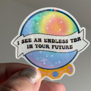 Könnte beinhalten: Ein holografischer Aufkleber mit einem Regenbogenhintergrund und Sternen. Der Aufkleber zeigt eine Wahrsagerkugel mit dem Text "I see an endless TBR in your future".