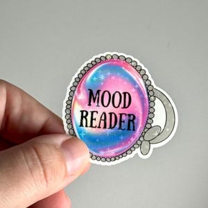 Könnte beinhalten: Ein Aufkleber mit einem Kristallkugel-Design. Die Kristallkugel hat ein rosa, blaues und weißes Wirbelmuster mit den Worten "MOOD READER" in Schwarz. Der Aufkleber hat einen silbernen Perlenrand und einen grauen Griff.