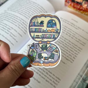 May include: A whimsical sticker featuring a witchy scene with a bookshelf, potion bottles, and pumpkins. The sticker has a holographic border and is held over an open book. The design includes a witch's cauldron and a crescent moon.