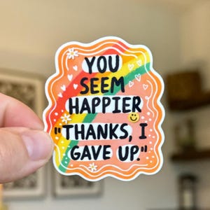 May include: A colorful sticker with the text "YOU SEEM HAPPIER THANKS, I GAVE UP." The design features a rainbow, hearts, and flowers. The sticker has an orange border and a smiling face.