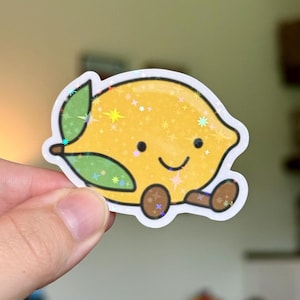 May include: A holographic sticker featuring a yellow lemon character with a smiling face, two small green leaves on the side, and two brown feet. The surface of the sticker has a sparkling, star-shaped iridescent finish that reflects light.