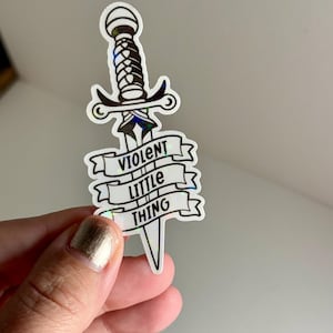 May include: A holographic sticker with a black and white illustration of a dagger with the text "VIOLENT LITTLE THING" on a banner.
