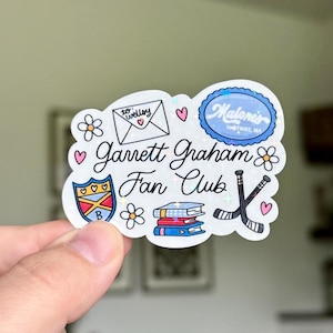 Peut inclure: Un autocollant blanc avec une bordure holographique avec le texte "Garrett Graham Fan Club". Le design comprend une lettre, des cœurs, des fleurs, un bouclier, des livres et une crosse de hockey. Le texte "Malone's Hastings, MA" est également inclus.
