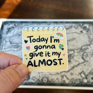 May include: A yellow notepad sticker with the text "Today I'm gonna give it my ALMOST." The sticker has a rainbow, hearts, and other colorful graphics. The sticker is held by a person's hand.