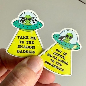 Könnte beinhalten: Zwei gelbe und grüne Aufkleber mit Alien-Motiv. Der erste Aufkleber zeigt einen Außerirdischen in einem fliegenden Untertasse mit dem Text "Take me to the shadow daddies". Der zweite Aufkleber zeigt einen Außerirdischen in einem fliegenden Untertasse mit einem Buch und dem Text "Get in bestie, we're going to the bookstore".