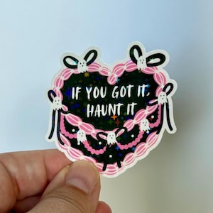 May include: A heart-shaped sticker with the text "IF YOU GOT IT, HAUNT IT." The sticker has a black background with a holographic effect, a pink scalloped border, and small ghost and bow illustrations.