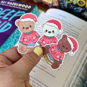 May include: Three Christmas bear stickers. Each bear wears a red sweater with a Christmas tree design and a Santa hat. The bears are brown, white, and dark brown. The stickers are held over an open book.