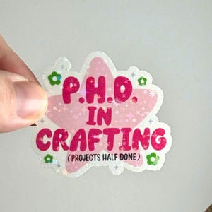 May include: A holographic sticker with a pink star shape and the words "P.H.D. IN CRAFTING" in pink. The sticker also includes the text "(PROJECTS HALF DONE)" and is decorated with small green flower and star accents.