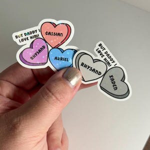 Puede incluir: Cinco pegatinas en forma de corazón con purpurina y texto. Las pegatinas dicen "But Daddy I Love Him!"  "Cassian", "Rhysand", "Azriel", y "Rhysand" y "Xaden".