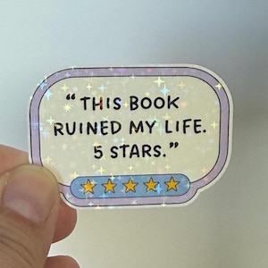 Może przedstawiać: Holograficzna naklejka z napisem "THIS BOOK RUINED MY LIFE. 5 STARS." Naklejka ma jasnofioletową obwódkę z gwiazdkami i rząd pięciu złotych gwiazd na dole.