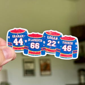 Könnte beinhalten: Ein gestanzter Aufkleber mit vier blauen Eishockeytrikots mit roten und gelben Verzierungen. Die Trikots zeigen die Namen GRAHAM 44, DI LAURENTIS 66, LOGAN 22 und TUCKER 46. Das Design ist mit einem holografischen Sternenmuster versehen.