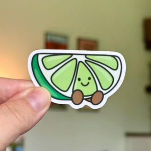 May include: A die-cut sticker shaped like a lime wedge with a light green interior, a dark green rind, and a white border. The lime wedge features a simple cartoon face with two black eyes and a small smile, along with two small brown feet.