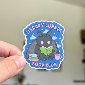 May include: A die-cut sticker featuring a fluffy dark grey moth character reading a green book. The background is blue with stars, clouds, and a crescent moon. The text reads LIBRARY LURKER BOOK CLUB in pink letters. A stack of books and a pink mug are included.