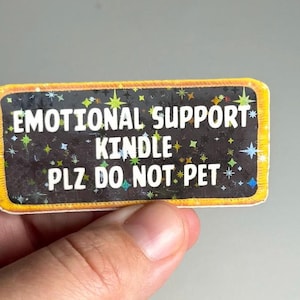 May include: A yellow and black rectangular sticker with a black background and white text that reads "Emotional Support Kindle - Plz Do Not Pet". The background is decorated with colorful stars.